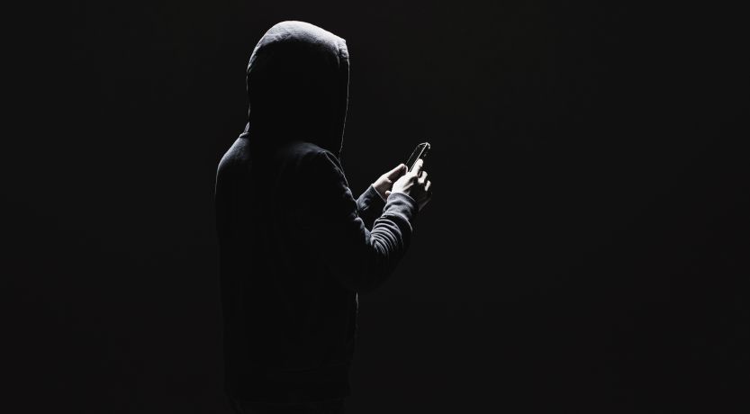 Dark photo of a man in a hoodie holding a phone to illustrate the article 