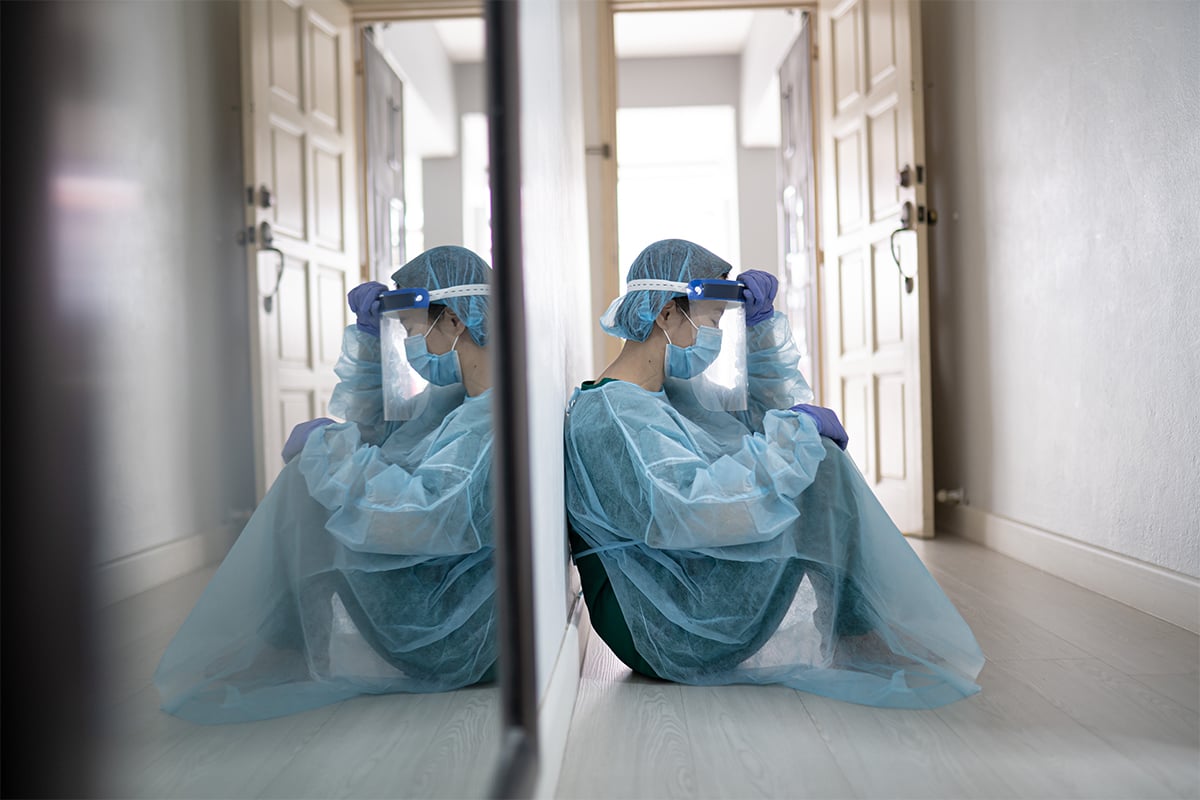 Photo of dejected health care worker in protective gear sitting on the floor to illustrate the article 