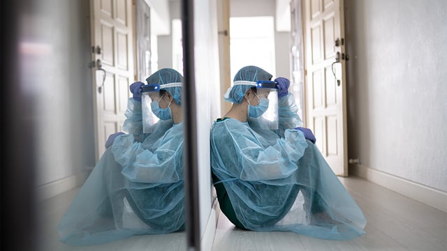 Photo of dejected health care worker in protective gear sitting on the floor to illustrate the article 